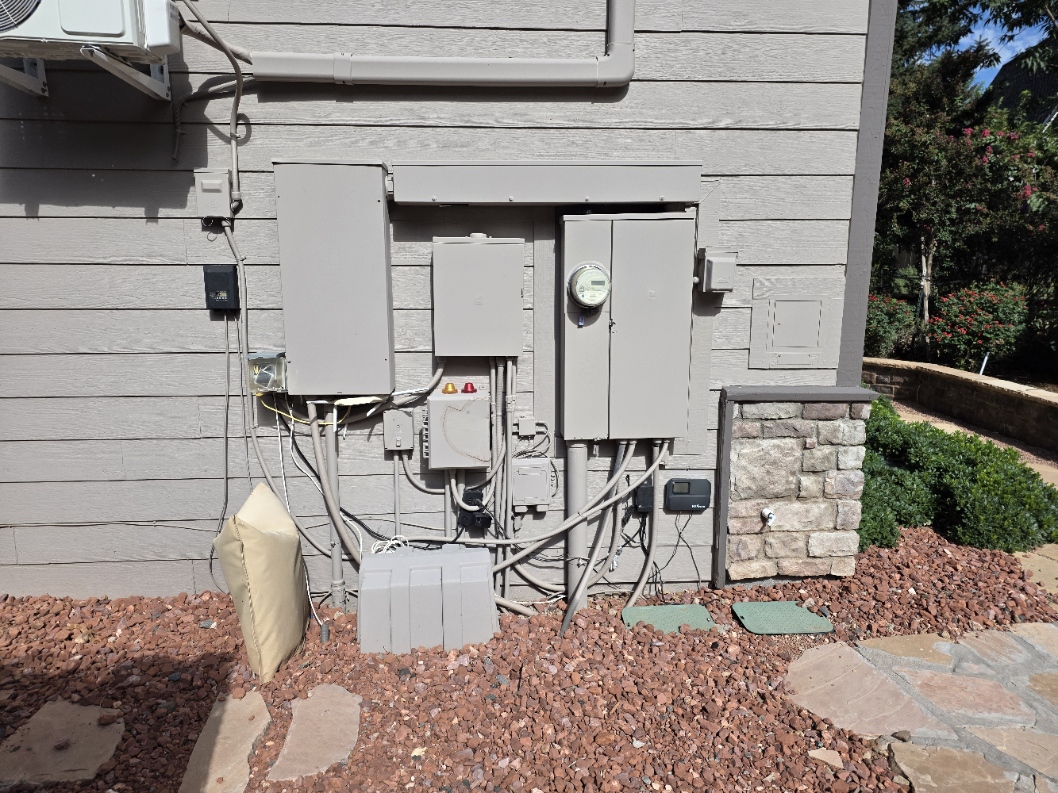 Exterior wall with two large electrical panels: panel 1 on the left for generator-supported essential circuits, and panel 2 on the right next to the meter for nonessential circuits. The generator itself is out of frame to the left.